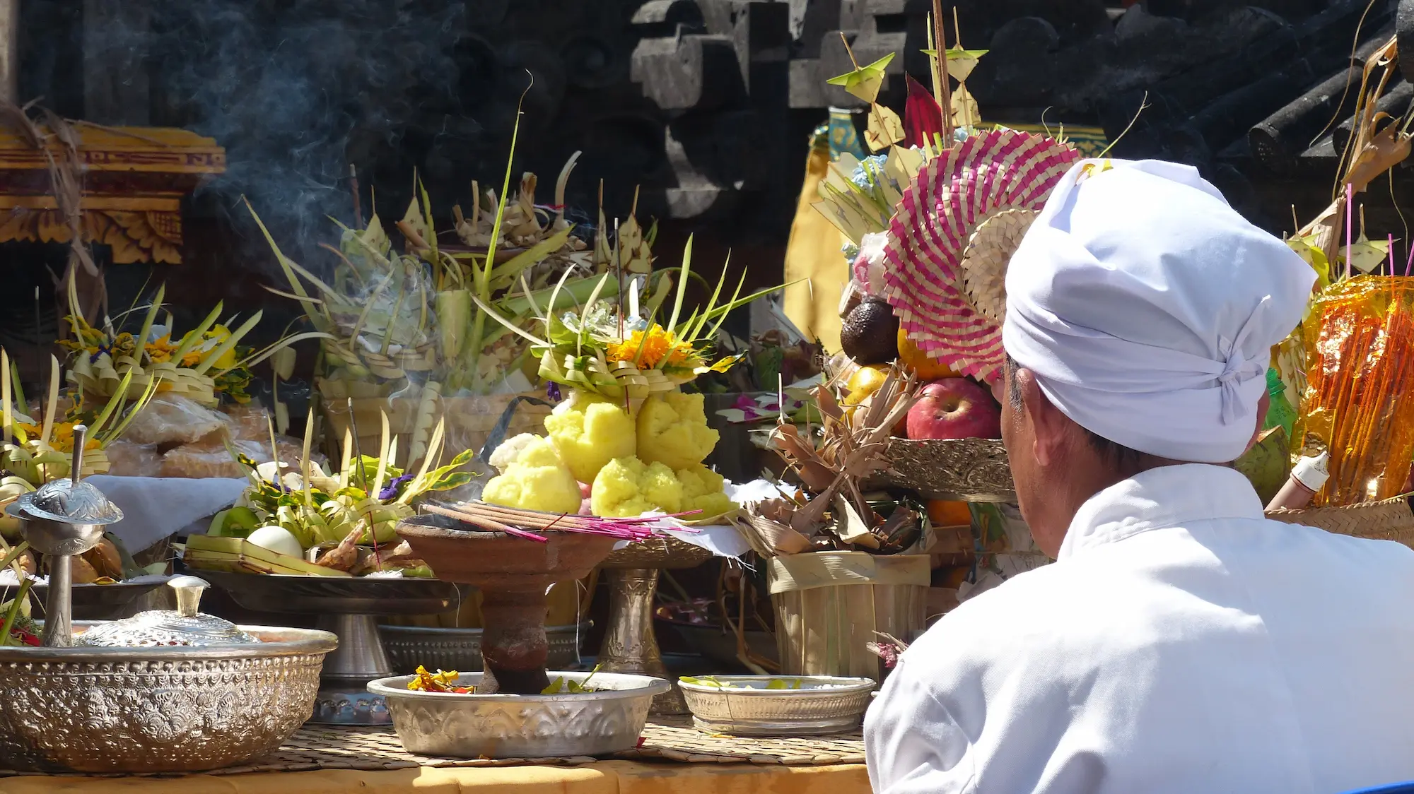 Hombre vestido de blanco ante un altar de ofrendas con humo de incienso en un templo de Bali